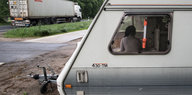 Blick durch das Fenster eines Wohnwagens auf eine sitzende Frau. Auf der Straße daneben ein Laster.