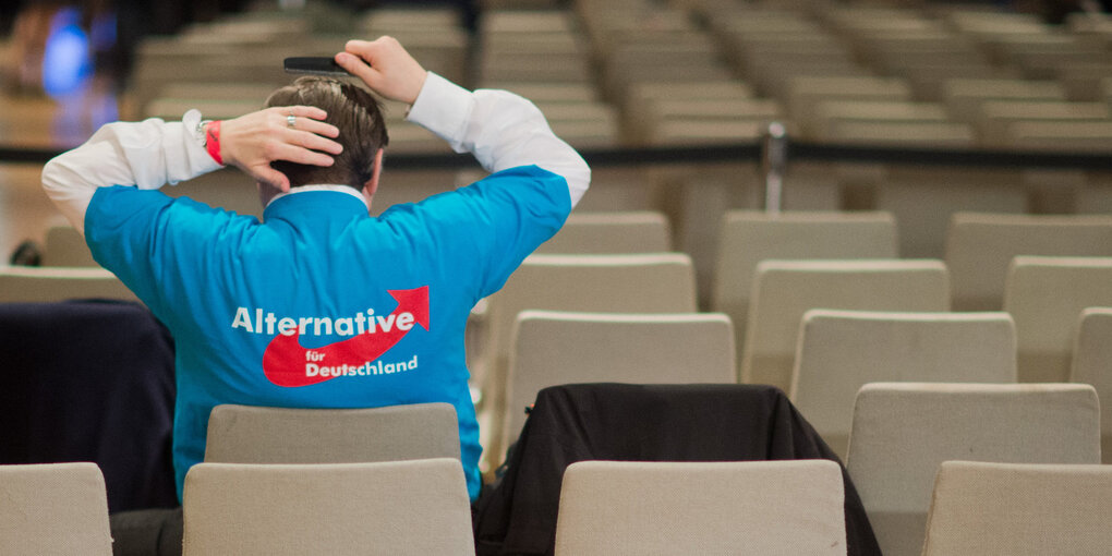 Mann im AfD-Shirt von hinten, im Hintergrund viele Stuhlreihen. Er kämmt sich die Haare.