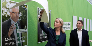 Eine Frau und ein Mann enthüllen Wahlplakate mit Winfried Kretschmann drauf, sie hängen an einer grünen Wand