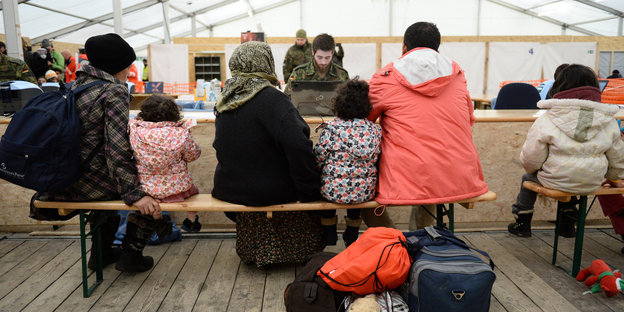 Mehrere Flüchtlinge mit Taschen sitzen mit dem Rücken zum Betrachter auf einer Bank in einem großen Zelt, vor ihnen sitzt ein Soldat vor einem Laptop
