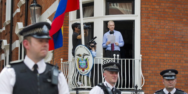 Julian Assange steht mit kurzen Haaren auf einem Hochparterre-Balkon, neben ihm die Flagge Ecuadors, vor ihm britsche Polizisten.