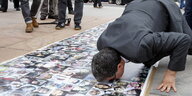 Mann kniet sich im Freien auf ein auf den Boden ausgebreitetes Banner mit Fotos nieder und küsst es.