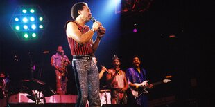 Mehrere Musiker von Earth, Wind & Fire stehen in unbschreiblichen Klamotten stehen auf einer Bühne und performen