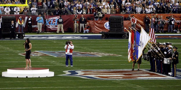 Eine Sängerin und Uniformierte mit Fahnen auf einem Football-Feld