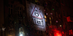 Ein Transparent mit der Aufschrift "1 KM" und einem Anarchiesymbol hängt an einem Gebäude