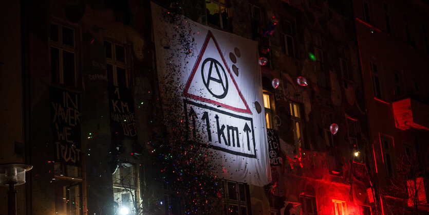 Ein Transparent mit der Aufschrift "1 KM" und einem Anarchiesymbol hängt an einem Gebäude