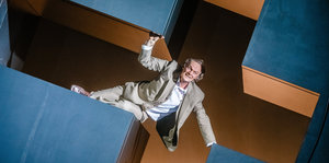 Ein Mann hängt schräg in einer kreuzförmigen Bühnendekoration und guckt wie Klaus Kinski