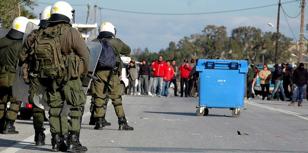 Polizisten stehen einer Gruppe Menschen gegenüber, zwischen ihnen steht ein blauer Müllcontainer