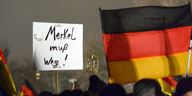 Demonstranten halten eine Deutschland-Flagge und ein Schild hoch. Auf dem Schild steht: "Frau Merkel muß weg!"