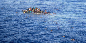 Ein Boot kentert, zahlreiche Menschen befinden sich noch auf ihm, einige schwimmen bereits im Wasser.