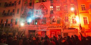 Dunkel gekleidete Menschen demonstriren auf der Straße. Die Häuser dahinter leuchten rot-orange und an den Häusern hängen Plakate.