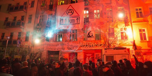 Dunkel gekleidete Menschen demonstriren auf der Straße. Die Häuser dahinter leuchten rot-orange und an den Häusern hängen Plakate.