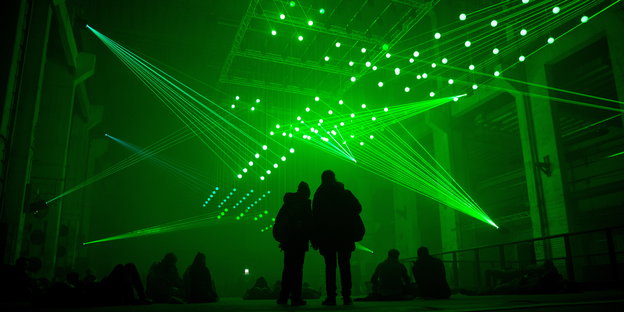 Menschen stehen und sitzen in einer großen Halle. Von der Decke strahlen viele Laser in den Raum.