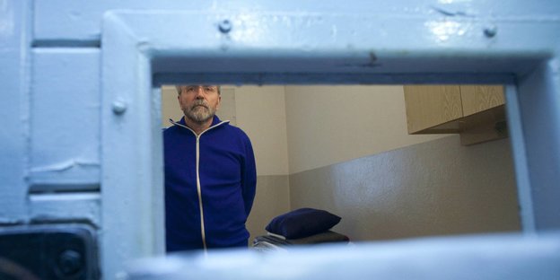 Blick durch ein Sichtklappe in eine Zelle hinein. darin steht ein Mann mit blauer Jacke.