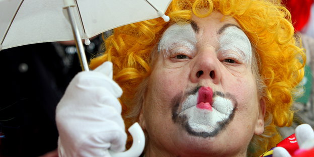 Zum Clown geschminkter Mann hält einen Regenschirm und wirft ein Küsschen und Richtung des Betrachters.