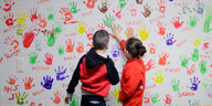 Zwei Kinder stehen vor einer Wand voller bunter Händeabdrücke
