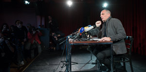 Yanis Varoufakis sitzt an einem Tisch vor einem Mikrofon, erleuchtet von Spots, sonst ist es dunkel