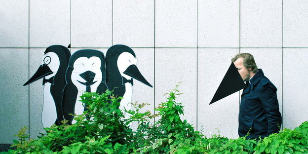 Etwas abseits von drei Pinguinen, die an eine Wand gemalt sind, steht ein Mann, der sich einen Schnabel aufgesetzt hat