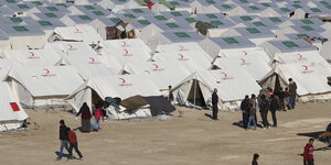 Viele weiße Zelte stehen auf sandigem Grund. Davor sind einige Menschen zu sehen.