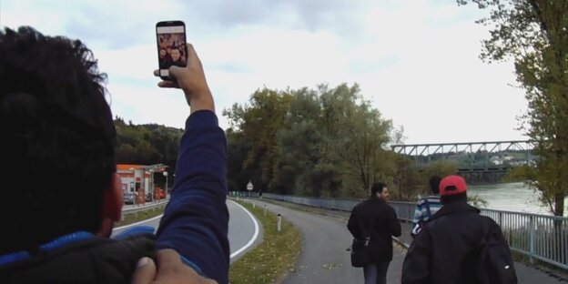 Ein Mensch macht ein Selfie von sich und anderen. Im Hintergrund ein Fluss und eine Brücke.