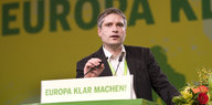 Sven Giegold steht an einem grünen Rednerpult mit einem Blumenstrauss in der Hand