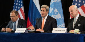 drei Männer sitzen vor Flaggen der UN, Russlands und der USA