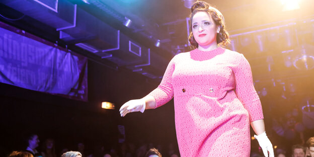 Ein Puls Size Model im pinken Kleid läuft über einen Laufsteg und lächelt