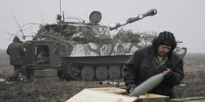 ein Soldat nimmt eine Granate aus einer Kiste, im Hintergrund ein Panzer