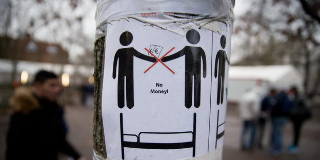 Ein Zettel klebt an einem Baum. Auf ihm sind zwei Personen zu sehen. Eine Person übergibt Geld, der Transfer ist durchgestrichen, darunter steht "No Money!"
