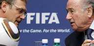 Zwei Männer stecken die Köpfe zusammen, sie reden vertraulich. Im Vordergrund ist ein Ball zu sehen, im Hintergrund steht Fifa.