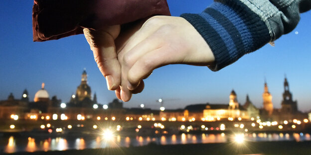Zwei Hände halten sich vor dem Hintergrund der Dresdner Silhouette am Abend.