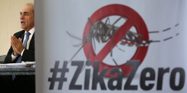 Ein Mann im Anzug hält seine Hände zusammengelegt hoch, neben ihm ein Schild, auf dem eine durchgestrichene Zika-Fliege und °'Zikozero" abgebildet ist