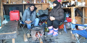 Ein Mann un eine Frau sitzen in einer provisorischen Bretterbehausung dick angezogen an einer kleinen Feuerstelle