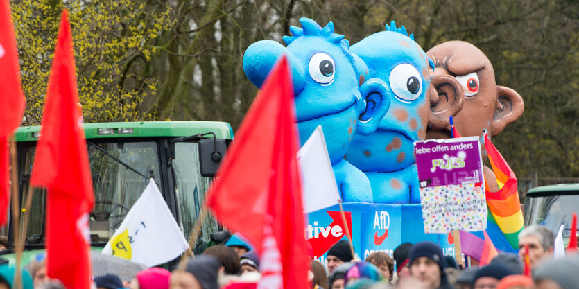 Ein Demoumzug trägt rote Fahnen und große drei große Puppen in blau, blau-braun und braun mich sich.