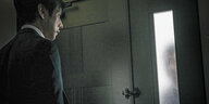 Ein Mann blickt auf einen Türspalt, hinter dem schemenhaft ein Schatten zu sehen ist.
