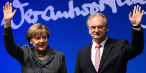 Angela Merkel und Reiner Haseloff stehen vor einem blauen Hintergrunf und winken in die Kameras.