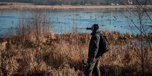 Ein Mann steht mit Fernglas in einer winterlichen Landschaft und beobachtet Vögel auf einem See.